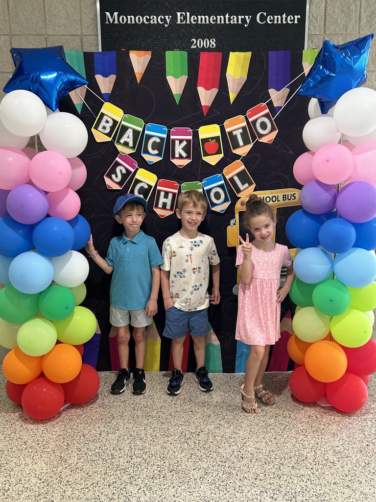 Three students standing in front of a black fabric background with a Back to School Banner, school bus and colored pencils. Each side of the background has rainbow colored balloon towers with a blue mylar star balloon on top.