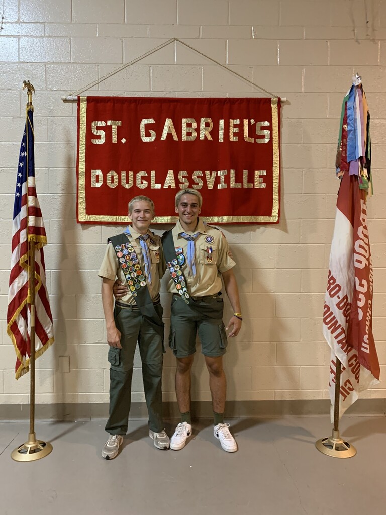Two scouting students wearing their uniforms with sashes standing in front of a St. Gabriel's Douglassville red and gold banner on a painted cement block wall. The American flag is on a pole on the left side of the photo and the boy scout Troop 595 flag is on a pole on the right side of the photo.