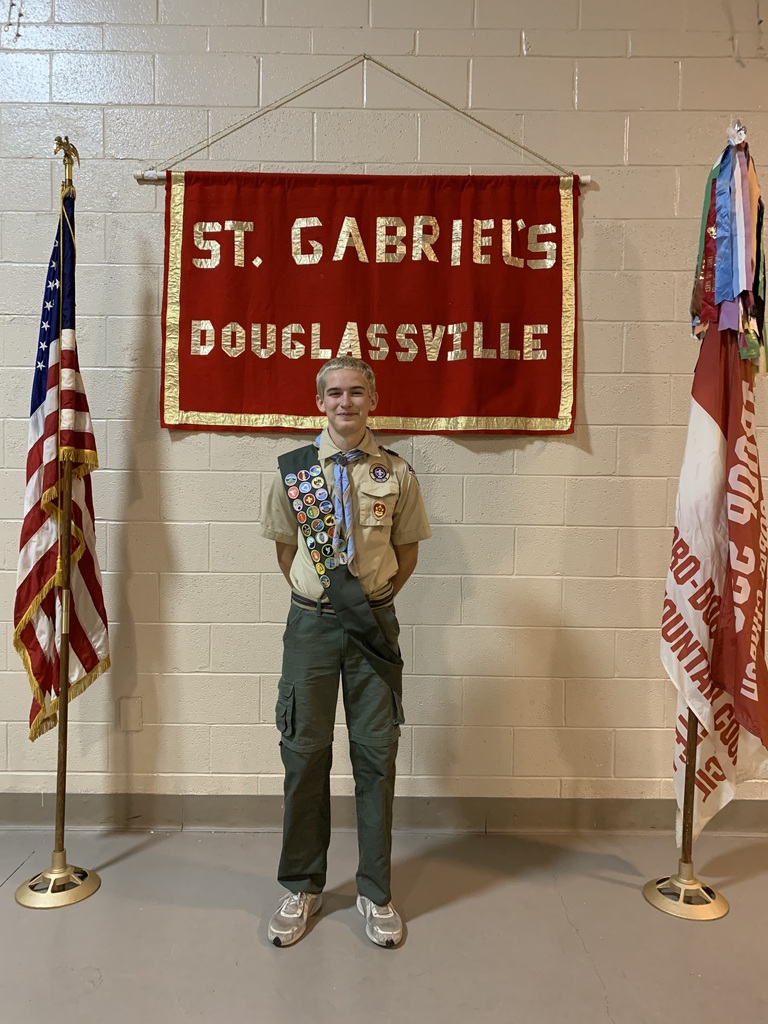 One scouting student wearing his uniform with sash standing in front of a St. Gabriel's Douglassville red and gold banner on a painted cement block wall. The American flag is on a pole on the left side of the photo and the boy scout Troop 595 flag is on a pole on the right side of the photo.