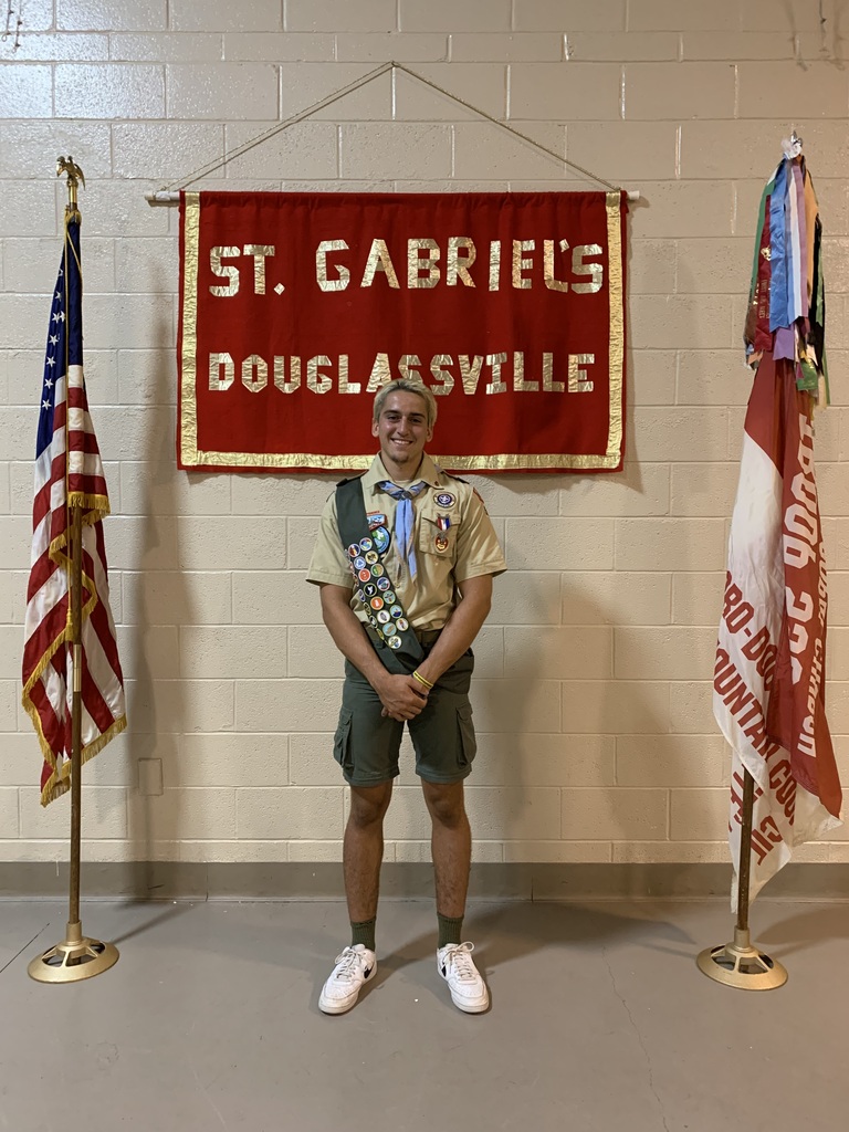 One scouting student wearing his uniform with sash standing in front of a St. Gabriel's Douglassville red and gold banner on a painted cement block wall. The American flag is on a pole on the left side of the photo and the boy scout Troop 595 flag is on a pole on the right side of the photo.