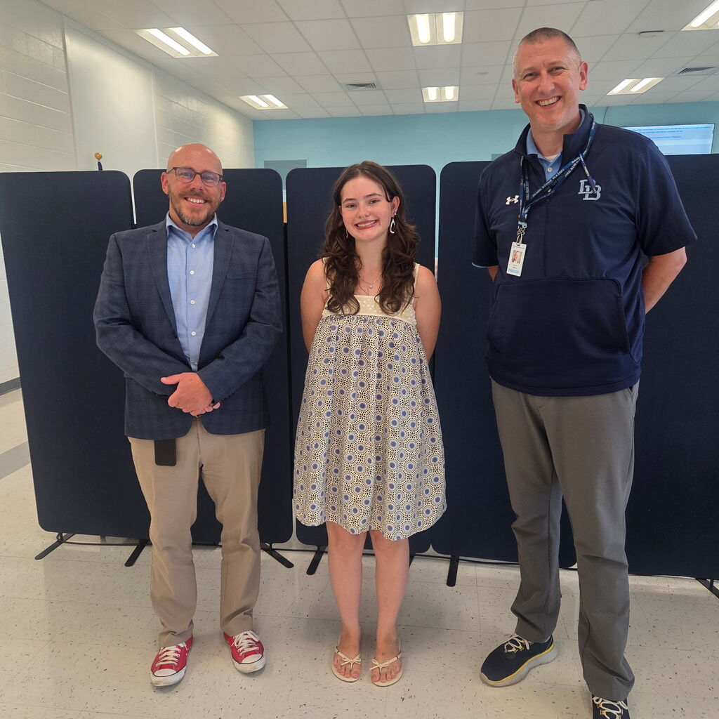 Three people stand side by side indoors for a recognition photo. In the center, Class of '26 grad Morgan Stevens wearing a sundress and sandals, recognizing her achievement of providing lifesaving measures to a family member. She is joined by Daniel Boone Area School District Superintendent Tom Voelker on the left and High School Principal Aaron Sborz on the right. All three are smiling while standing in front of black presentation boards in a bright school room.