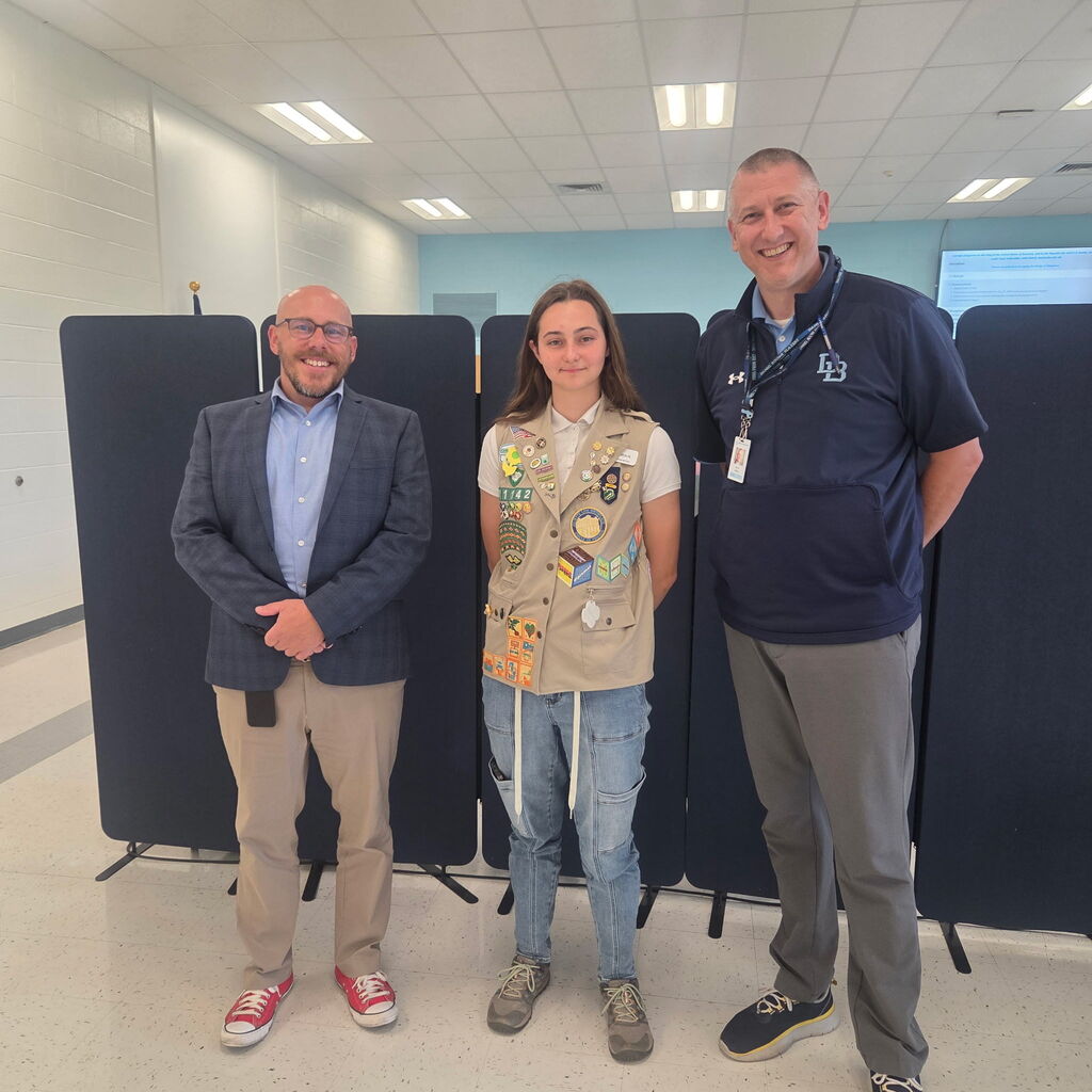 Three people stand side by side indoors for a recognition photo. In the center, Girl Scout Gwendolyn Harding wears her tan Girl Scout vest covered with badges and patches, recognizing her achievement of earning the Girl Scout Gold Award. She is joined by Daniel Boone Area School District Superintendent Tom Voelker on the left and High School Principal Aaron Sborz on the right. All three are smiling while standing in front of black presentation boards in a bright school room.
