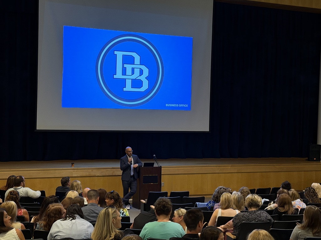 Superintendent Tom Voelker speaks to staff in the middle school auditorium. DB logo on screen with the word Business Office at the bottom. Tom stands behind a podium holding a microphone while staff sit in auditorium seats with their backs to the camera.