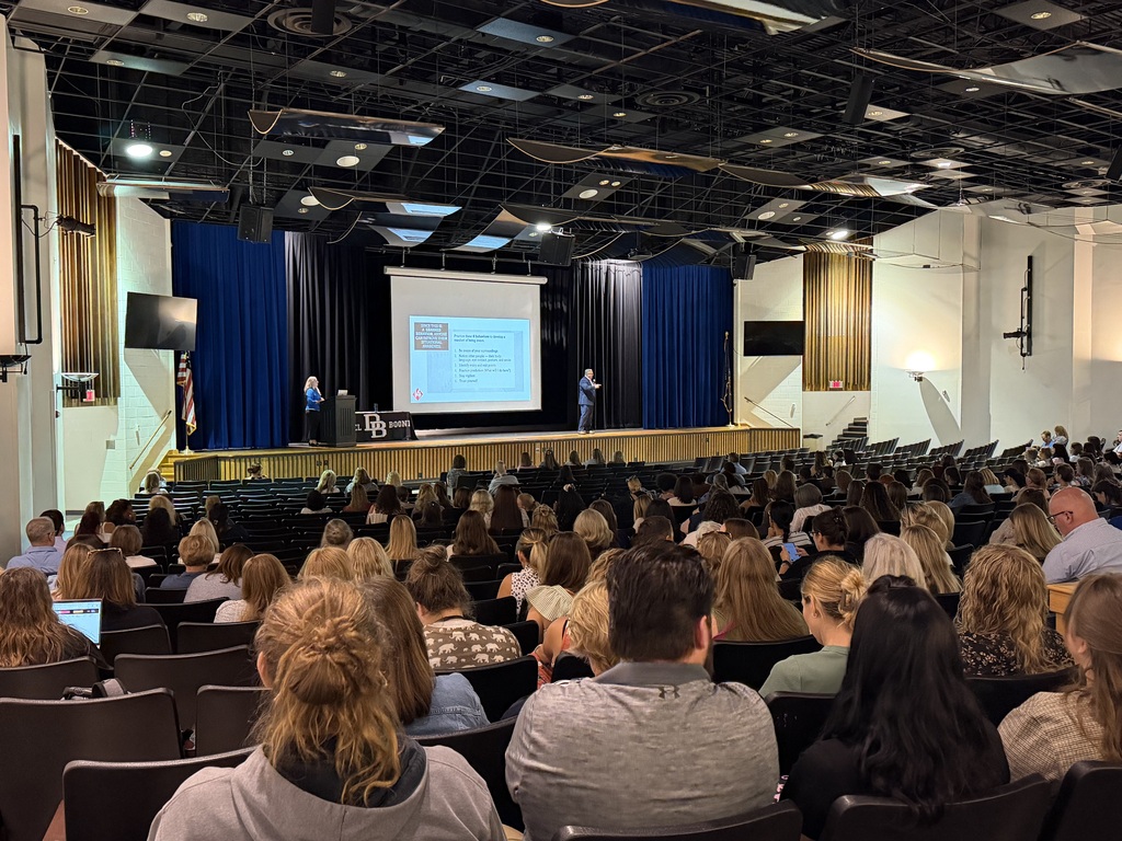 A large audience fills the Daniel Boone Area High School auditorium during an Opening Day Kick-Off event. Two presenters are on stage in front of a projection screen displaying a presentation, with Daniel Boone branding visible on the podium. Blue and black curtains frame the stage, while attendees sit throughout the auditorium listening to the presentation.