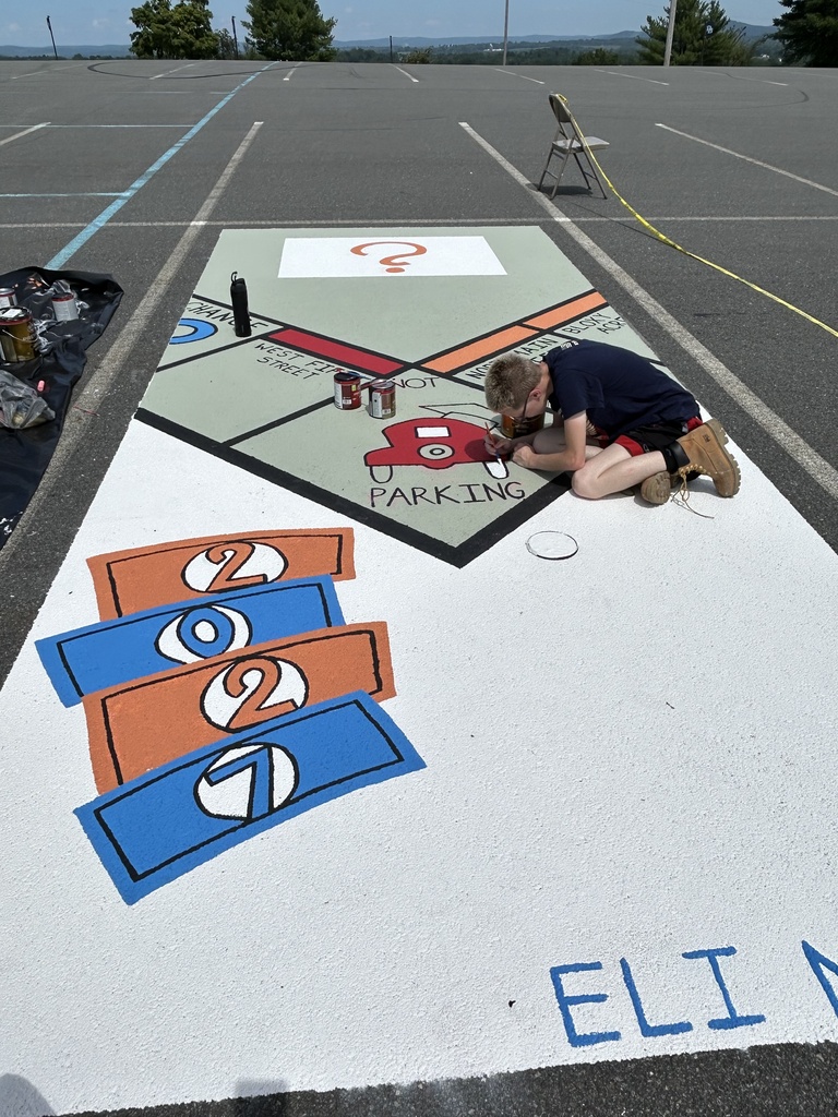 A student kneels while painting a parking spot decorated like a colorful Monopoly map, including street names, a red car-shaped parking symbol, and numbered game-like spaces in orange and blue.