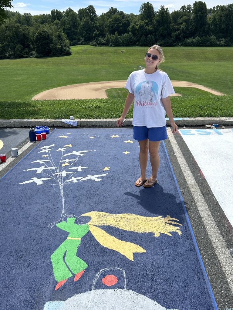 A senior stands beside a freshly painted parking spot featuring a whimsical blue background with a green character wearing a yellow scarf, surrounded by white flowers and yellow stars. A large yellow crescent moon fills the foreground.