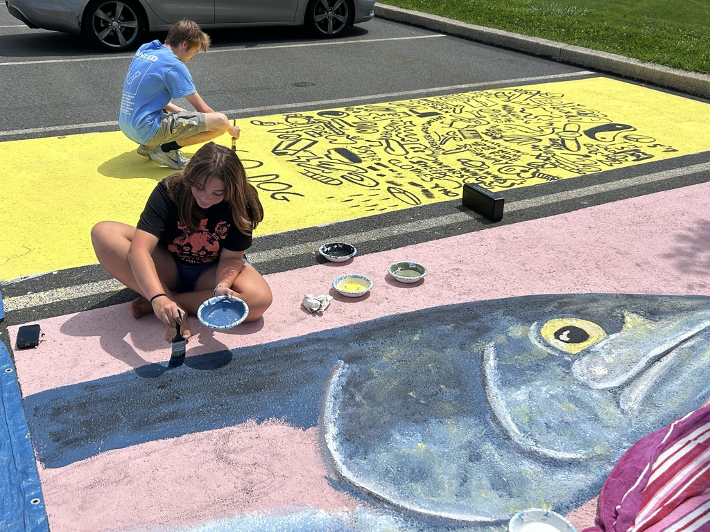 Two students work together painting a colorful parking spot. One student paints a large pink and blue fish design while another works on a bright yellow section covered with black doodles, drawings, and words.
