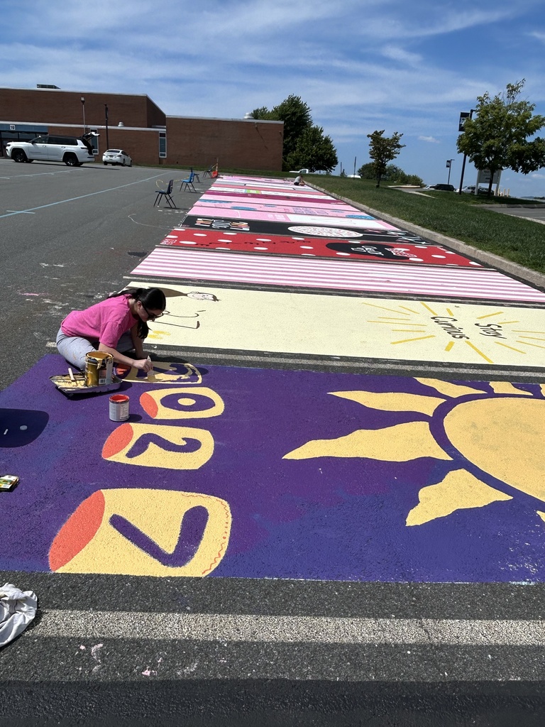 A student paints a large purple parking spot featuring oversized yellow and orange numbers and a large stylized yellow sun. Rows of brightly colored painted parking spots stretch into the distance along the high school lot.