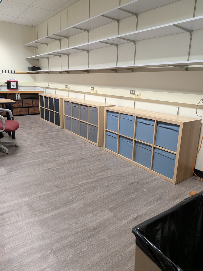 A classroom storage area with three wooden cubby units containing blue fabric bins, positioned beneath rows of wall-mounted shelves. Additional shelving, a desk, and a chair are visible in the background.
