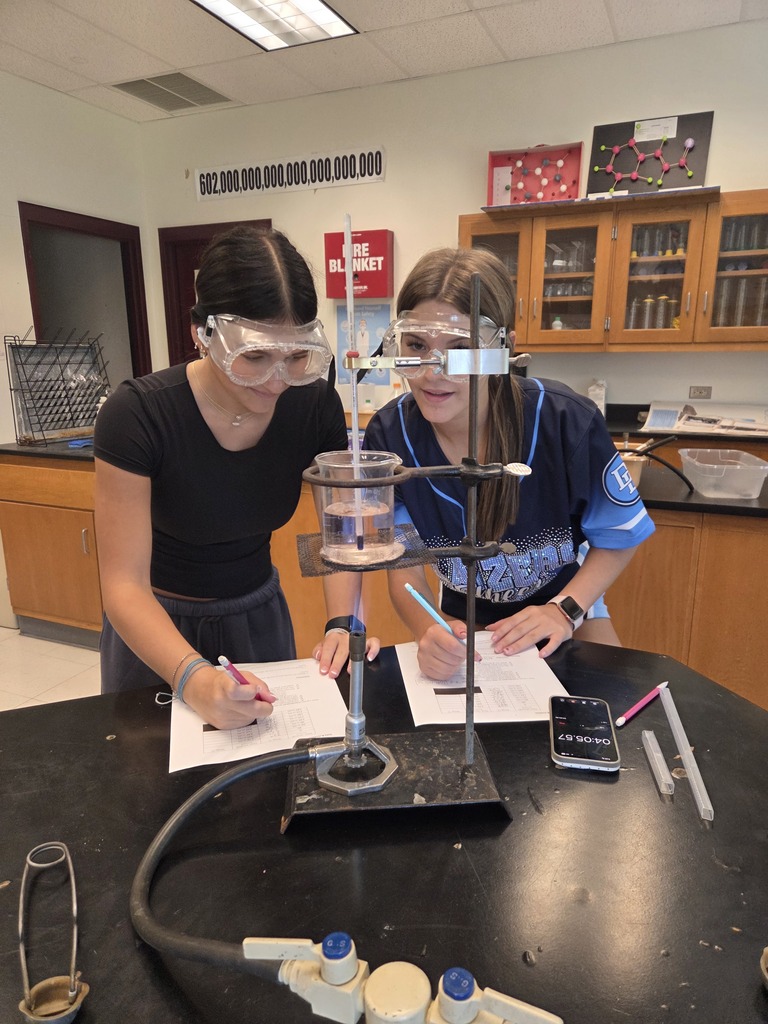 Two students in a chemistry lab wear safety goggles while recording data from a beaker of water being heated over a Bunsen burner. A thermometer is suspended in the beaker, and both students are writing on lab worksheets. Chemistry models and classroom cabinets are visible in the background.