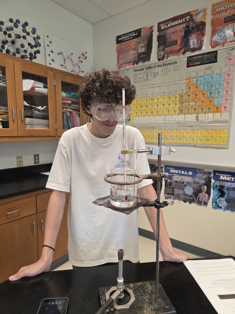 A student wearing safety goggles stands beside a laboratory setup with a beaker of water, thermometer, and Bunsen burner. The student observes the experiment while a worksheet rests on the lab table. A periodic table and chemistry posters are visible in the background.