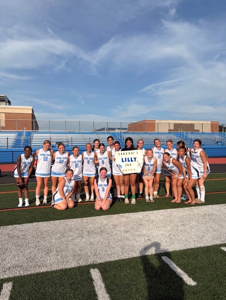Daniel Boone field hockey players pose together on the field with senior Lilly Maskeri holding a sign celebrating her 200 saves milestone. The team stands in front of the blue bleachers on a sunny day.