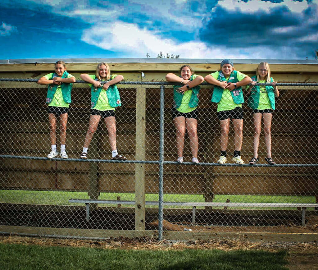 Five girl scouts wearing lime green tshirts, black shorts and green Girl Scout vests stand on a chain link fence in front of the new dugout they built