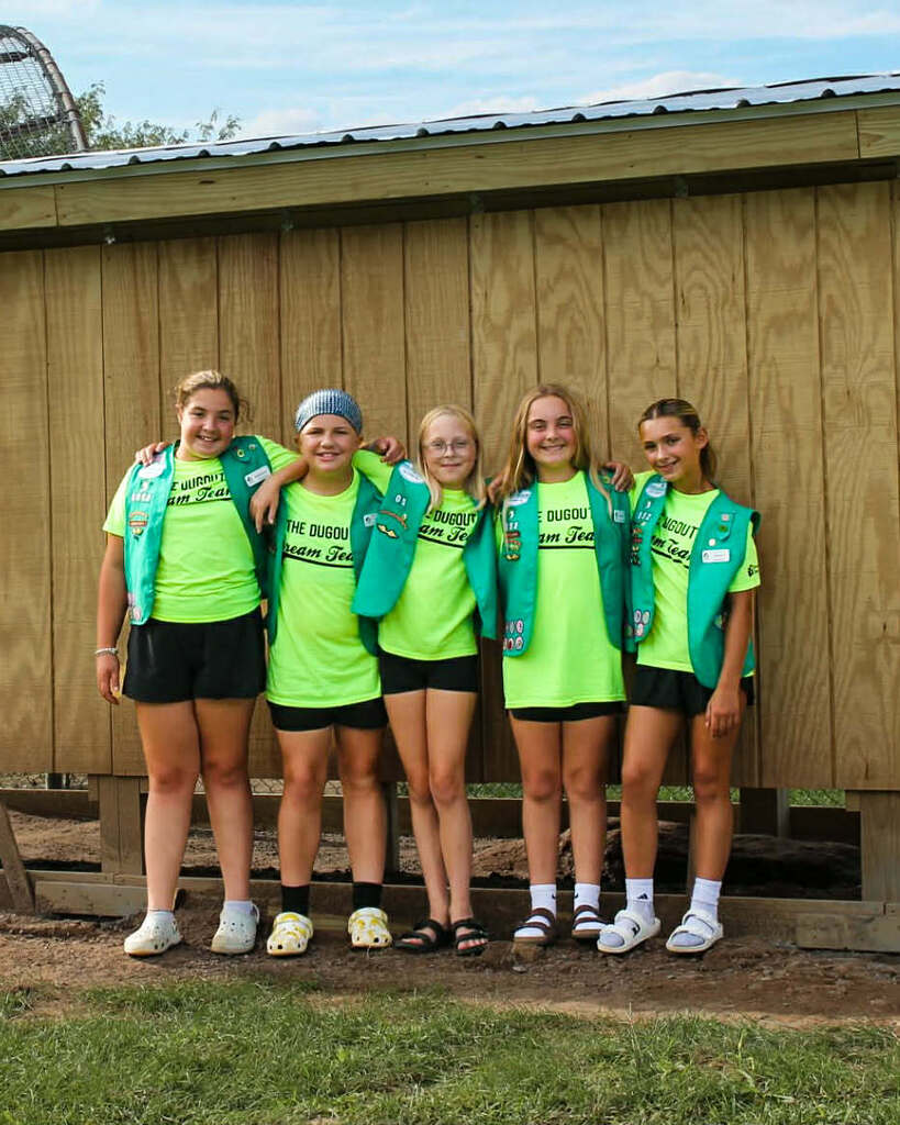 Five girl scouts wearing fluorescent green tshirts, black shorts and their green girl scout vests stand arm in arm in front of the dugout they built