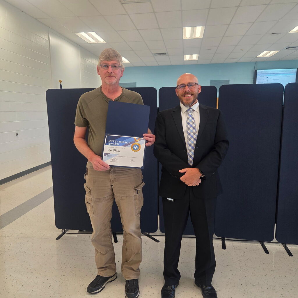 Superintendent Tom Voelker stands beside Sweet Impact Award recipient Kenny Templin. Both are smiling and standing in front of a partition in the board room.