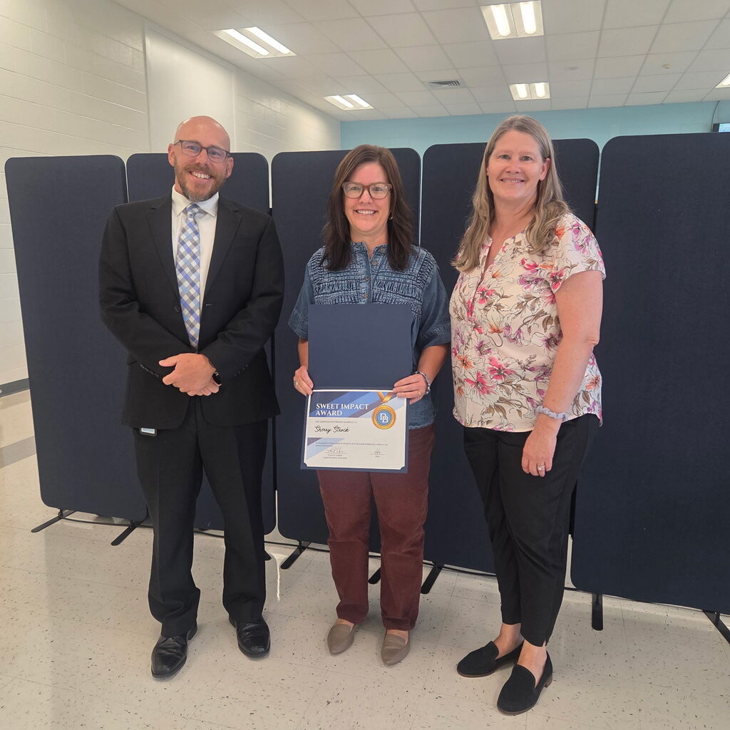 Superintendent Tom Voelker stands beside Sweet Impact Award recipient Sherry Strock. Primary Center Principal Melanie Hefter stands on the other side of Sherry. All are smiling and standing in front of a partition in the board room.