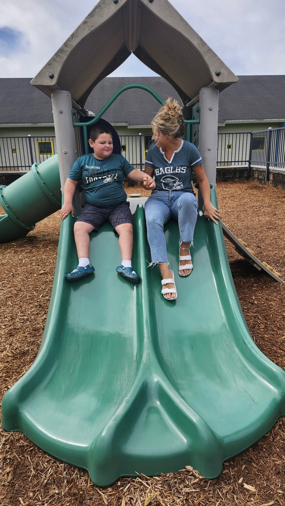 Student and teacher each wearing Eagles shirts slide down a dual slide together while holding hands.