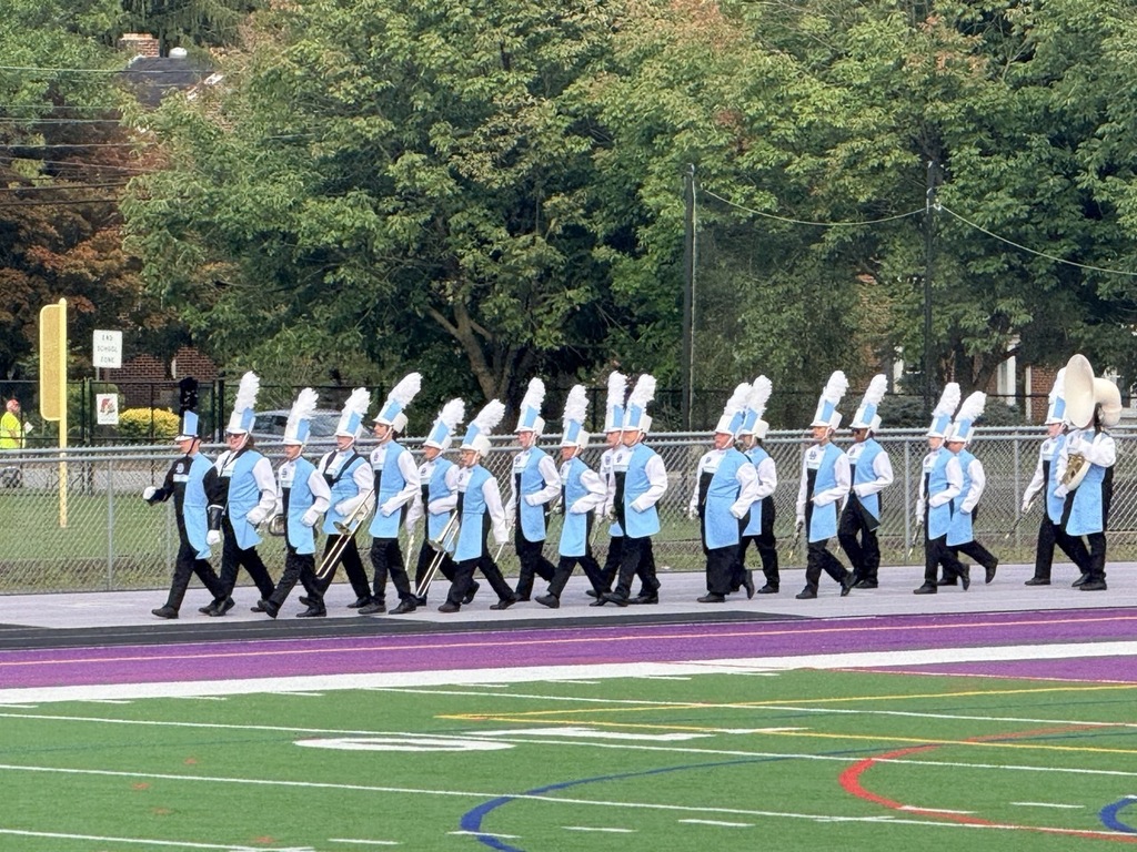 Daniel Boone Marching Blazers members in Carolina blue and white uniforms walk in double file on the track of a football field carrying their instruments.