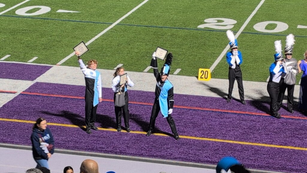 Daniel Boone Marching Blazers members in Carolina blue and white uniforms holding their framed certificates in the air after winning. All are standing on the track portion of a school football field.