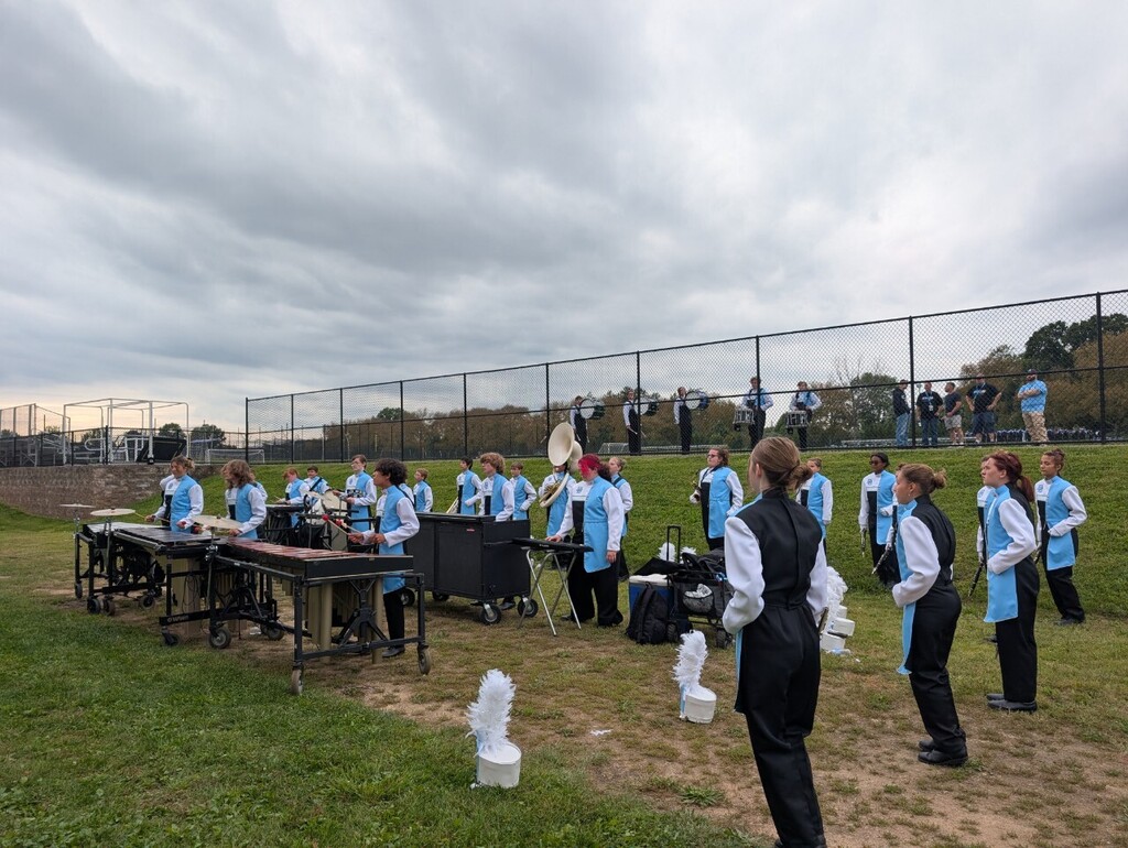 Daniel Boone Marching Blazers members in Carolina blue and white uniforms gather on a grassy field with percussion instruments and sousaphones before a performance. Color guard members stand in the foreground, with additional musicians and spectators visible in the background.