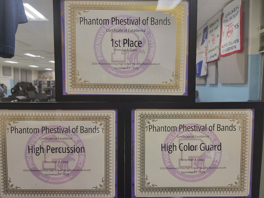 Photo of framed certificates stacked in a classroom window display. Each of the three certificates are framed in black frame and have the heading Phantom Phestival of Bands with a gold decorative border on the certificate. One is 1st Place America A Class, one is High Percussion and the last is High Color Guard.