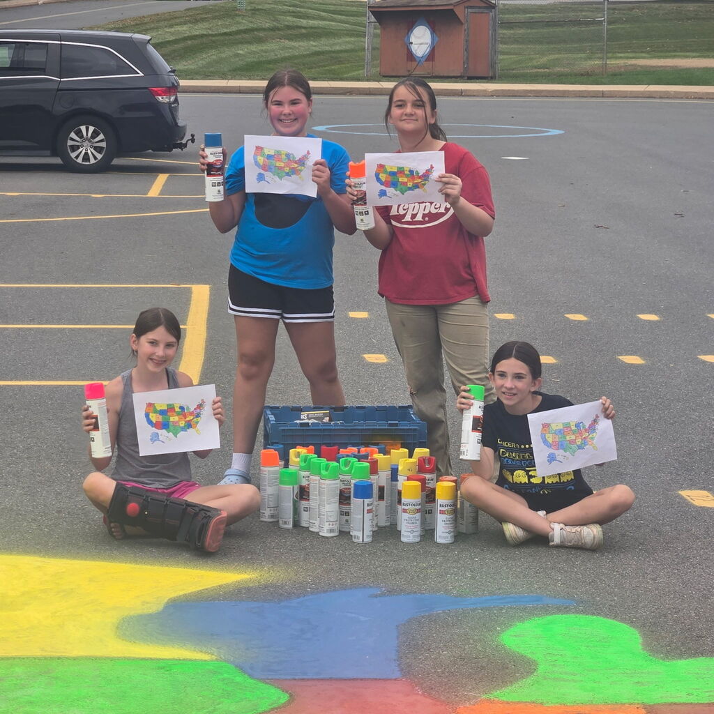 Four students holding colorful printed map page of the US with cans of fluorescent spray paint. Two girls are sitting on the playground and two girls are standing.
