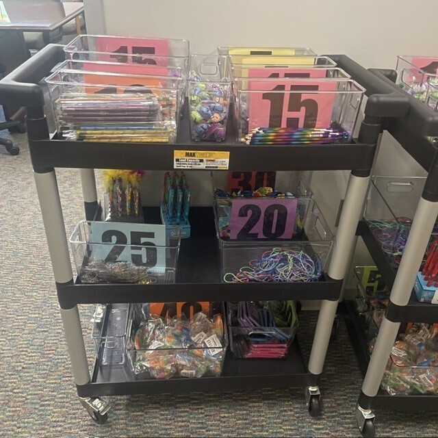 Wheeled three shelf cart with clear bins of prizes such as pencils, pens, fidgets and more.