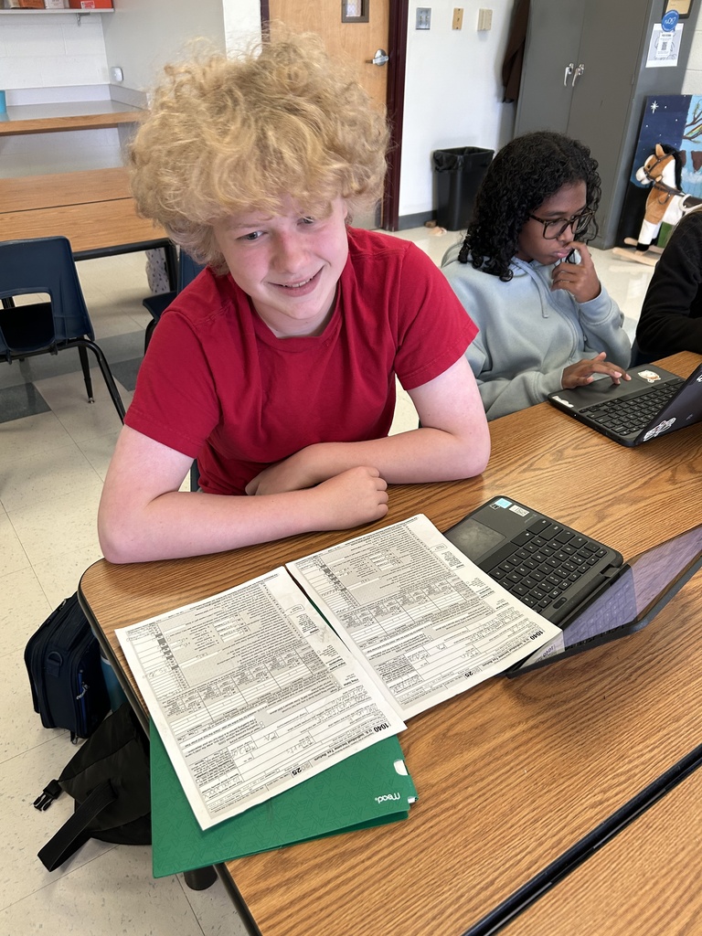 A high school student works on a tax return at a classroom table while a classmate works on a laptop nearby. Completed tax forms are spread across the table.