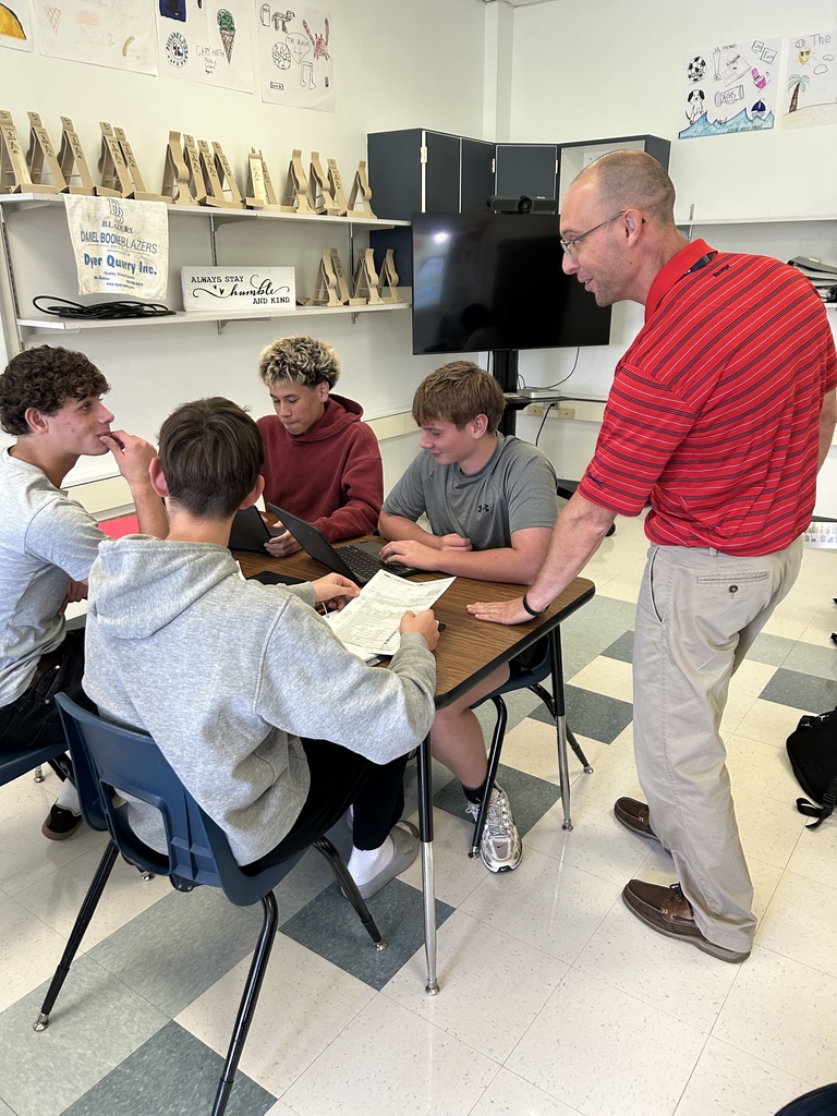 Mr. Stamy works with four high school students seated around a table as they review tax forms on laptops and paper documents during a Personal Finance class.