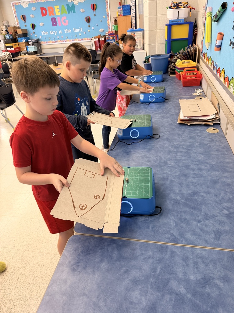 Four students stand at a counter holding cardboard pieces they are using Chompsaws to cut shapes they drew on the cardboard.