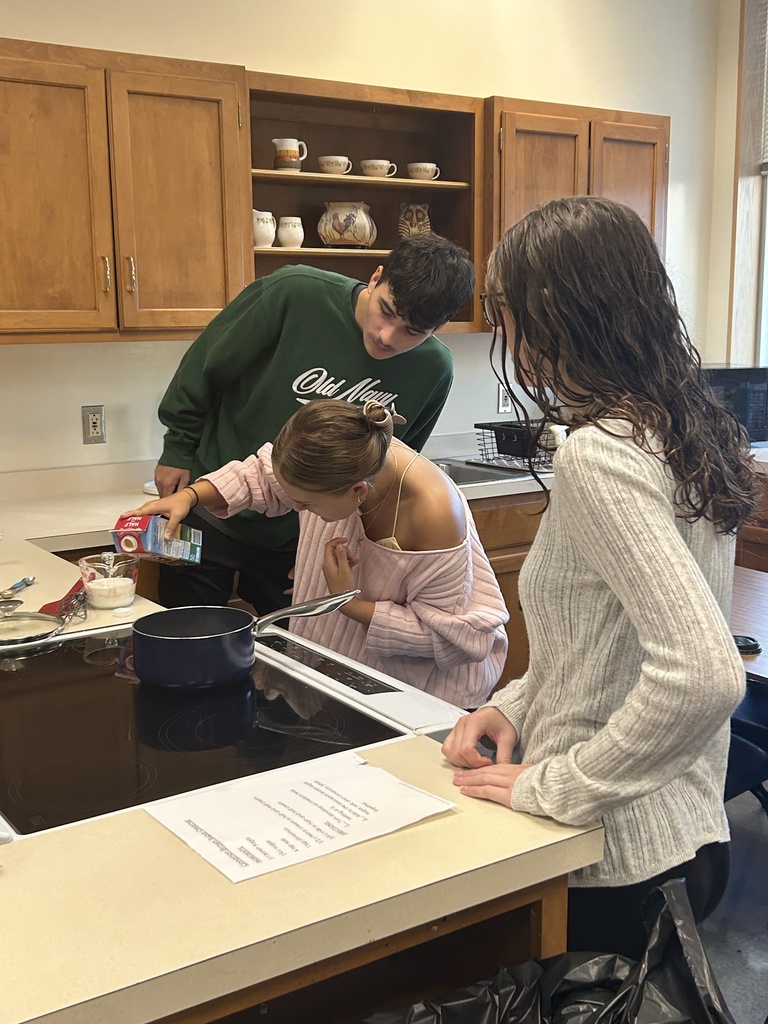 Three students stand at a counter. Two watch as one measures half and half into a pyrex mixing cup.
