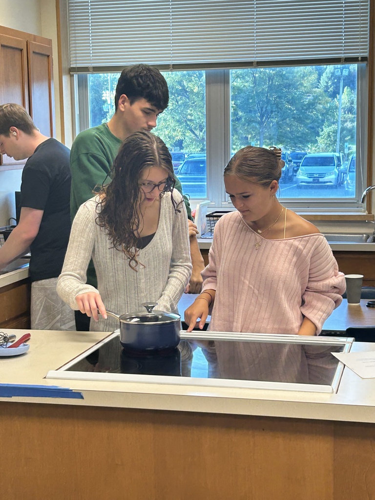 Two students are adjusting the heat under a pot on a stovetop.