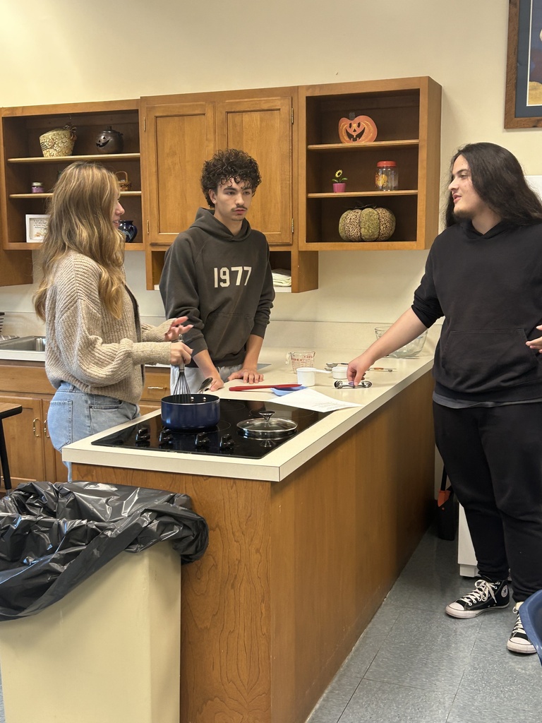 Three students stand by a stovetop. One is whisking the ingredients in a pot while the others discuss what's next.