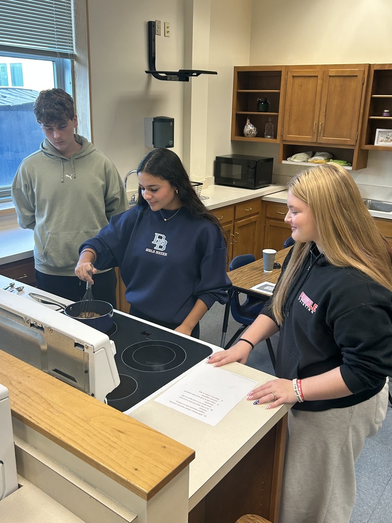 Three students stand by a stove. Two are watching while one whisks the ingredients of a pot on one of the burners.
