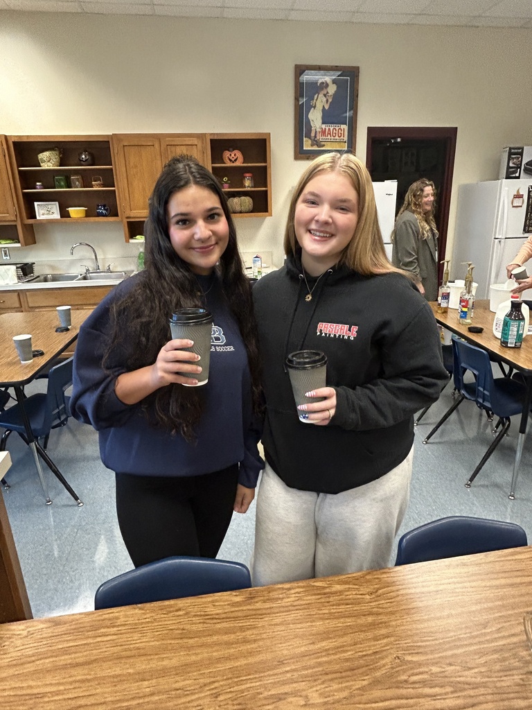 Two students stand holding disposable coffee cups with black lids. They are smiling at the camera.