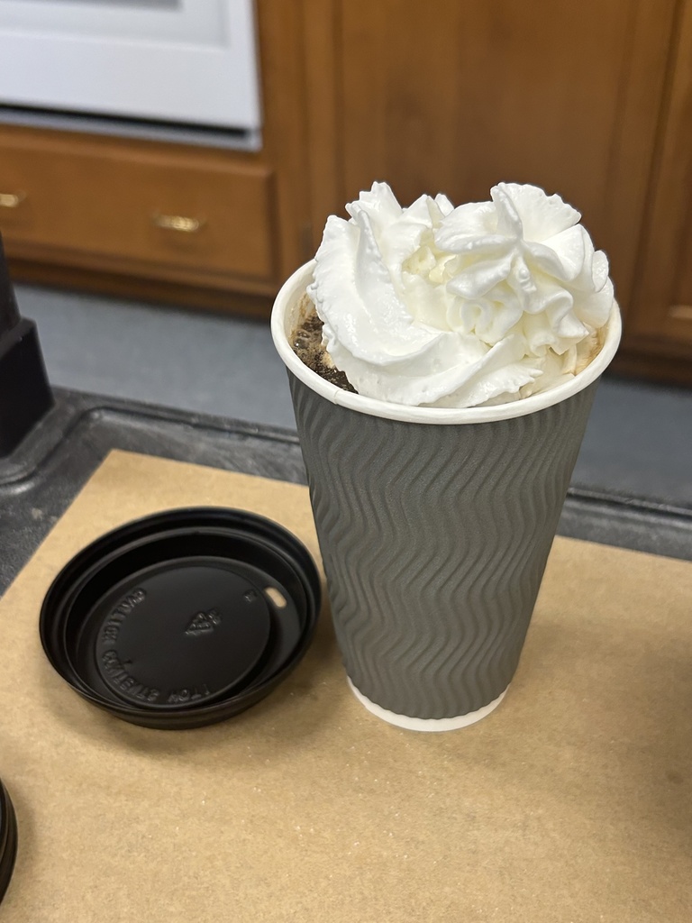 The finished product...a grey disposable coffee cup filled with iced coffee and topped with whipped cream. The black lid sits next to the disposable cup.