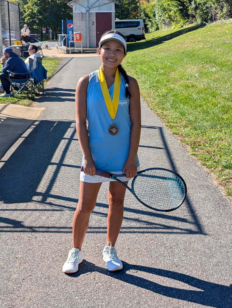 Student wearing Blazer blue tank and white skort and sneakers holding a tennis racquet wearing a yellow ribbon with bronze medal around her neck.