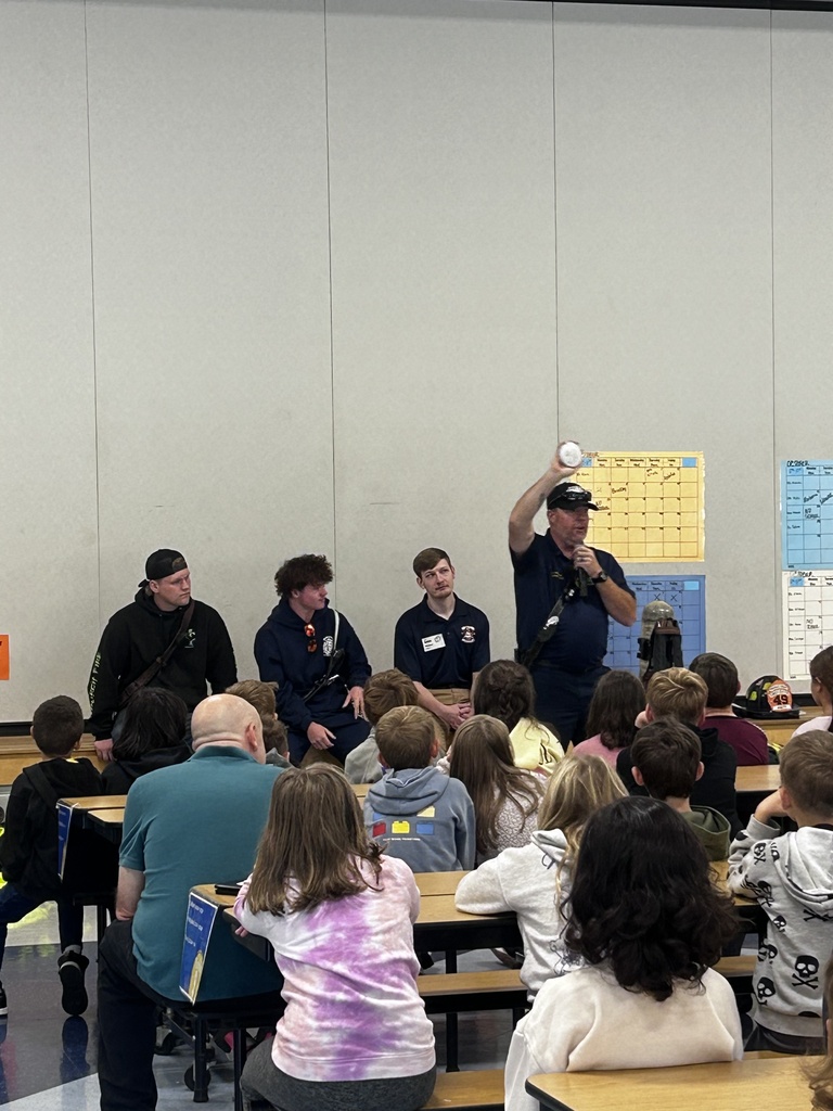 Firefighter holds a smoke detector while talking to the students about fire safety. Students are sitting at cafeteria tables watching the presentation.