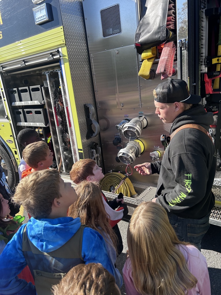 Students listening to a firefighter while they look at the equipment on the fire truck outside on the playground.