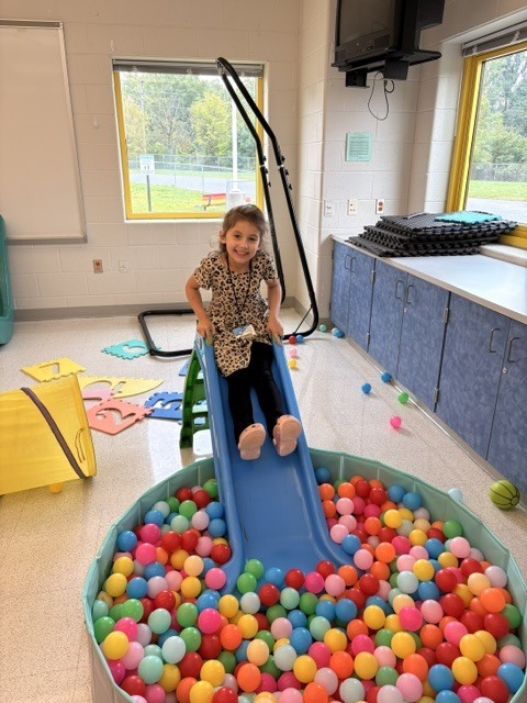 Student sitting on a child size plastic slide about to slide into a pit of colorful plastic balls.
