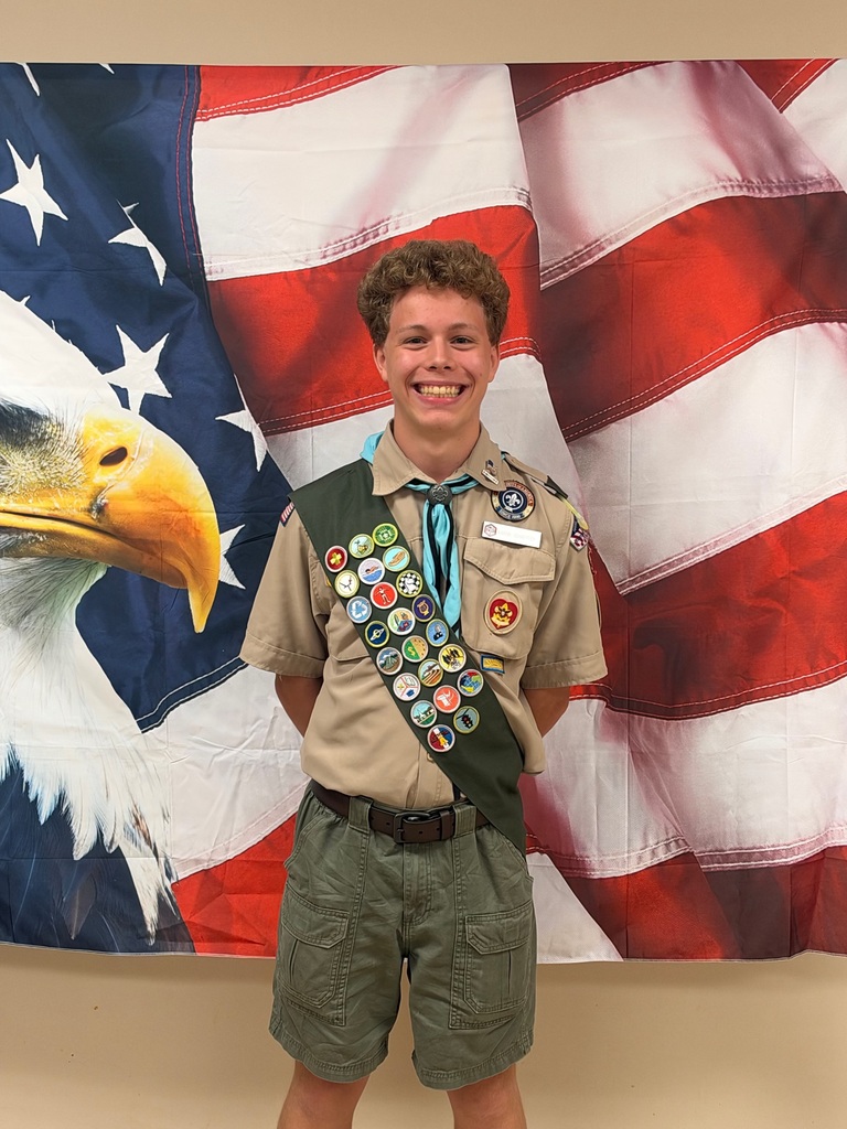 Eagle Scout stands in front of an American flag with a large eagle on the left side in front of the flag. The Eagle Scout is wearing his uniform and sash with badges.