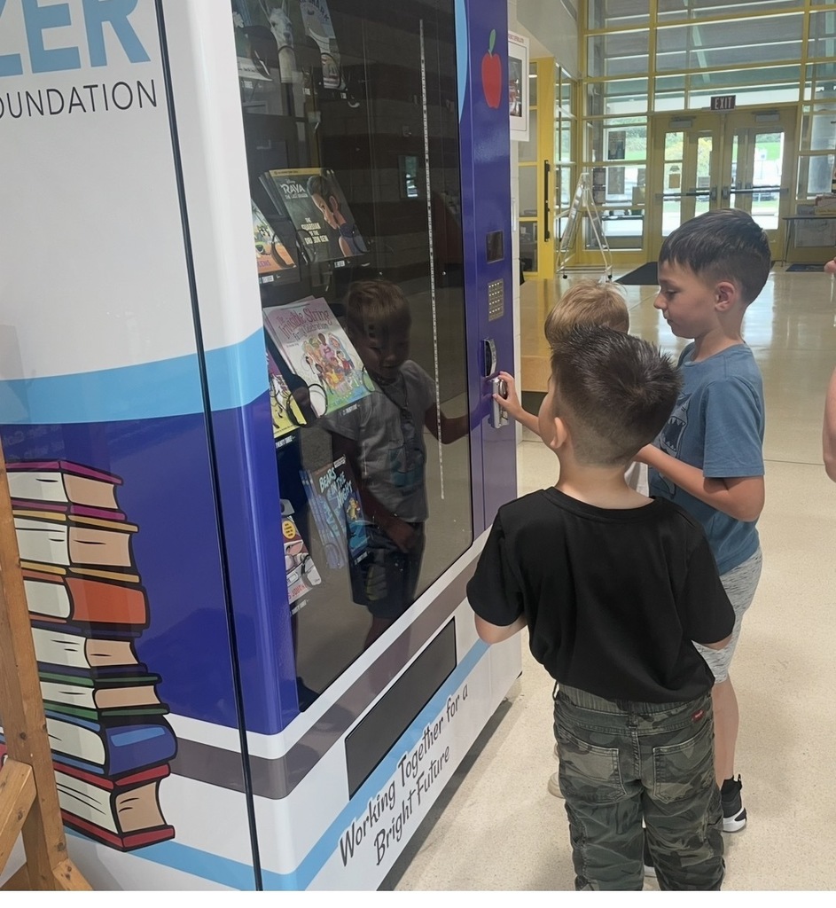 Three students stand in front of the Blazer Education Foundation Book Vending Machine as one student turns the handle to select a book.