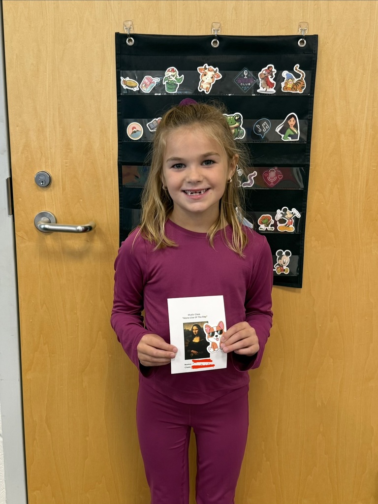 A smiling student holds their Mona Lisa of the Day award in Music class, standing in front of a classroom door decorated with character stickers.