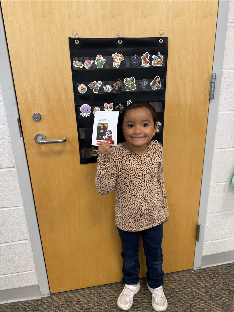 A smiling student holds their Mona Lisa of the Day award in Music class, standing in front of a classroom door decorated with character stickers.