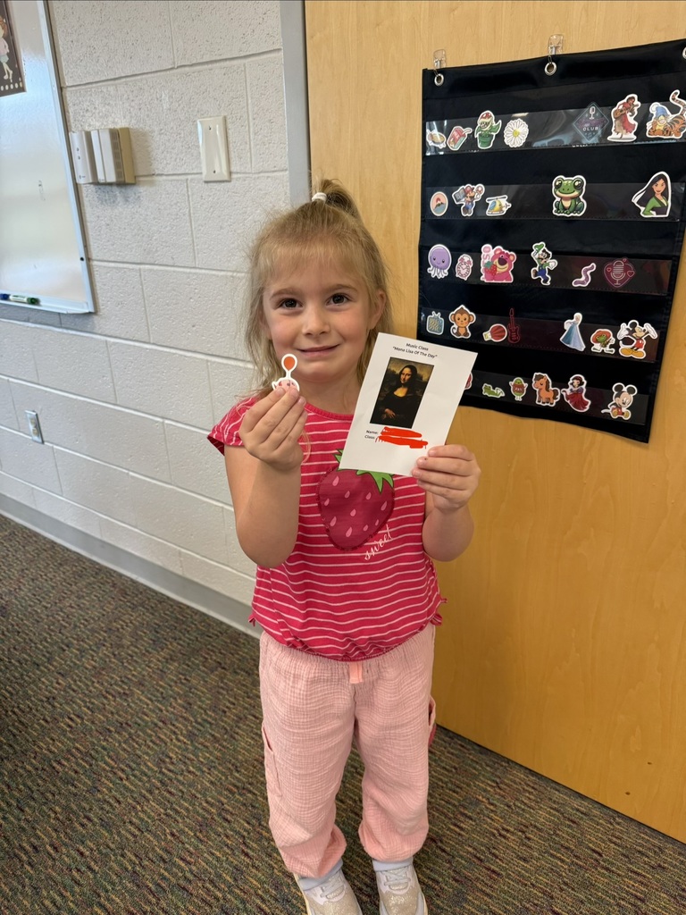A smiling student holds their Mona Lisa of the Day award in Music class, standing in front of a classroom door decorated with character stickers.