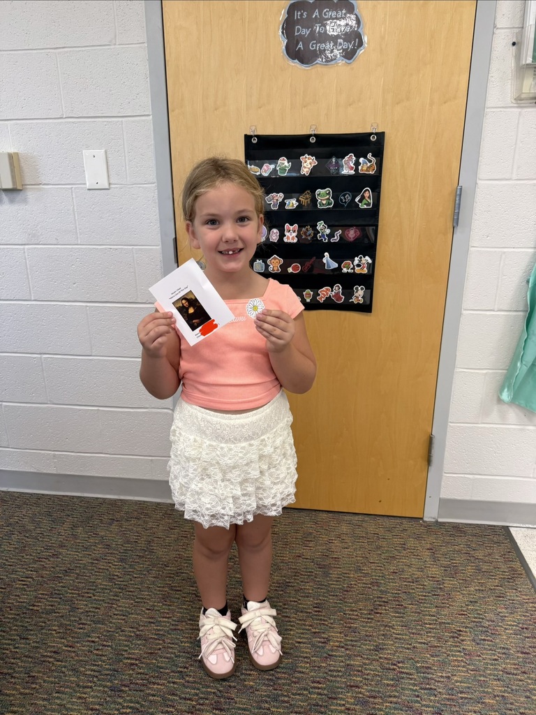 A smiling student holds their Mona Lisa of the Day award in Music class, standing in front of a classroom door decorated with character stickers.