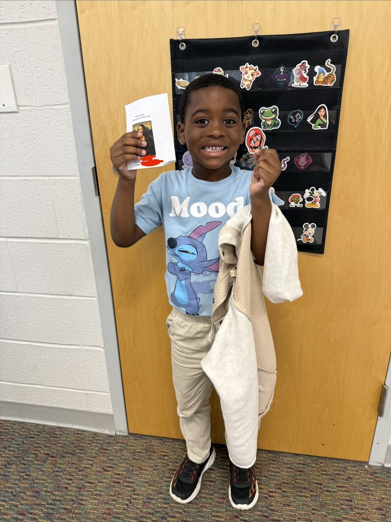 A smiling student holds their Mona Lisa of the Day award in Music class, standing in front of a classroom door decorated with character stickers.