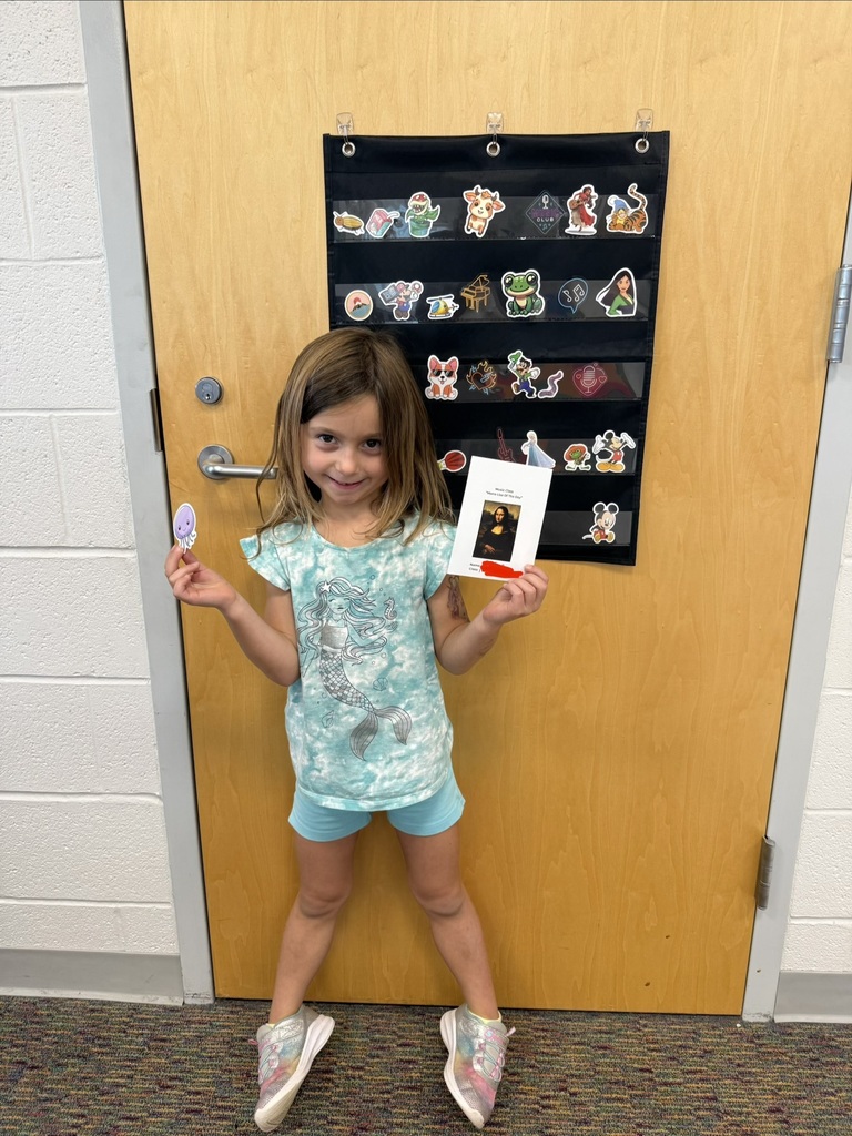 A smiling student holds their Mona Lisa of the Day award in Music class, standing in front of a classroom door decorated with character stickers.