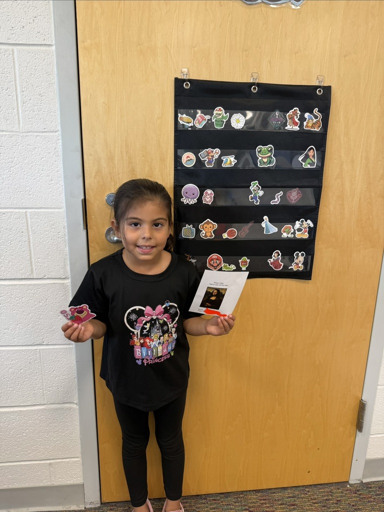 A smiling student holds their Mona Lisa of the Day award in Music class, standing in front of a classroom door decorated with character stickers.