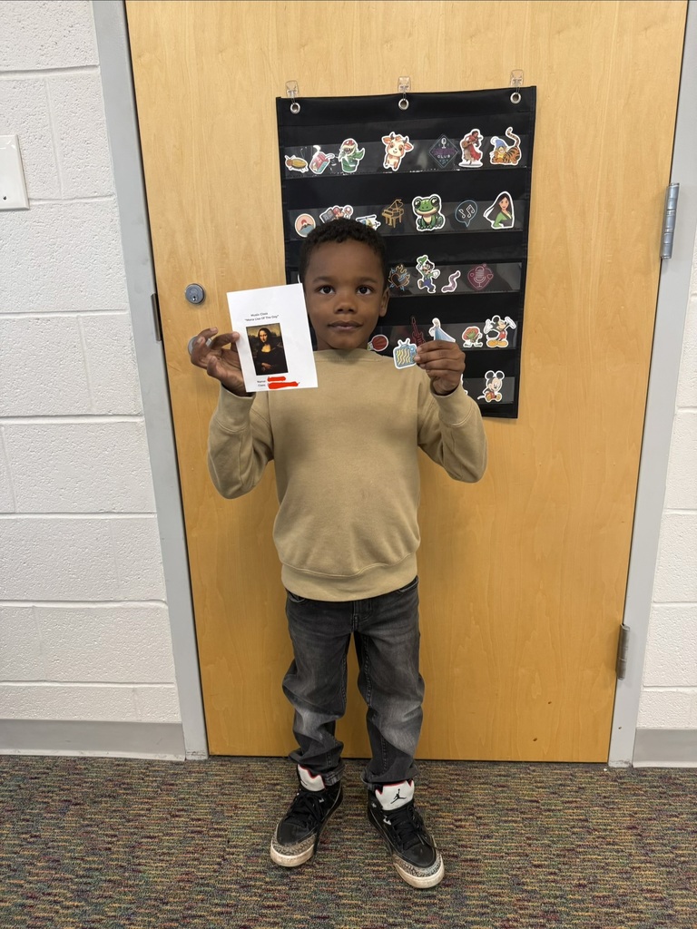A smiling student holds their Mona Lisa of the Day award in Music class, standing in front of a classroom door decorated with character stickers.